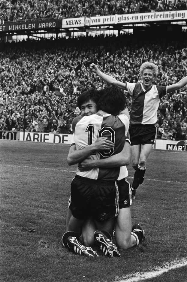 Drei Fußballspieler feiern ein Tor auf dem Boden, mit jubelnder Menge und schwenkenden Fahnen im Hintergrund, in Schwarz-Weiß abgebildet.