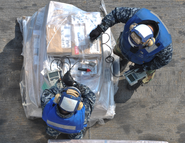 Zwei Männer in blauen Uniformen und Helmen arbeiten an einem großen Drohnen, halten verschiedene Gegenstände und haben eine Plastikabdeckung vor sich auf dem Boden.
