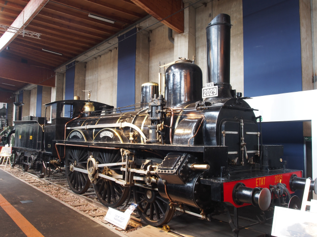 Alte Dampflokomotive in einem Museum mit umliegenden Eisenbahnschienen, Informationsschildern und anderen Ausstellungen, vor einer Wand mit Deckenleuchten.