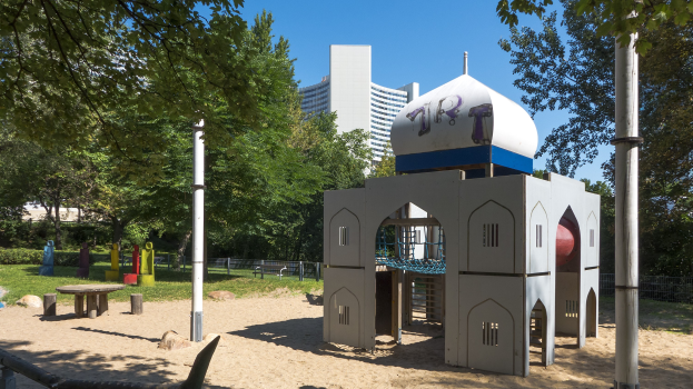 Spielplatz mit einem zentralen Spielgerüt, umgeben von Bäumen, Pfosten, Bänken und Gebäuden unter einem klaren blauen Himmel.