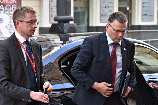 Zwei elegant gekleidete Männer im Anzug und Krawatte, beide mit Brille, steigen aus einem Auto aus, wobei einer eine Mappe hält, vor einem Gebäude mit Glastüren und Texttafeln.