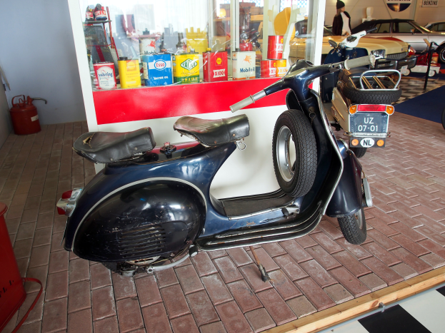 Ein schwarzer und weißer Roller steht vor einer Tankstelle, links daneben ein rotes Objekt und ein Feuerlöscher sowie mehrere Fahrzeuge, eine Person und an der Wand angebrachte Schilder im Hintergrund.