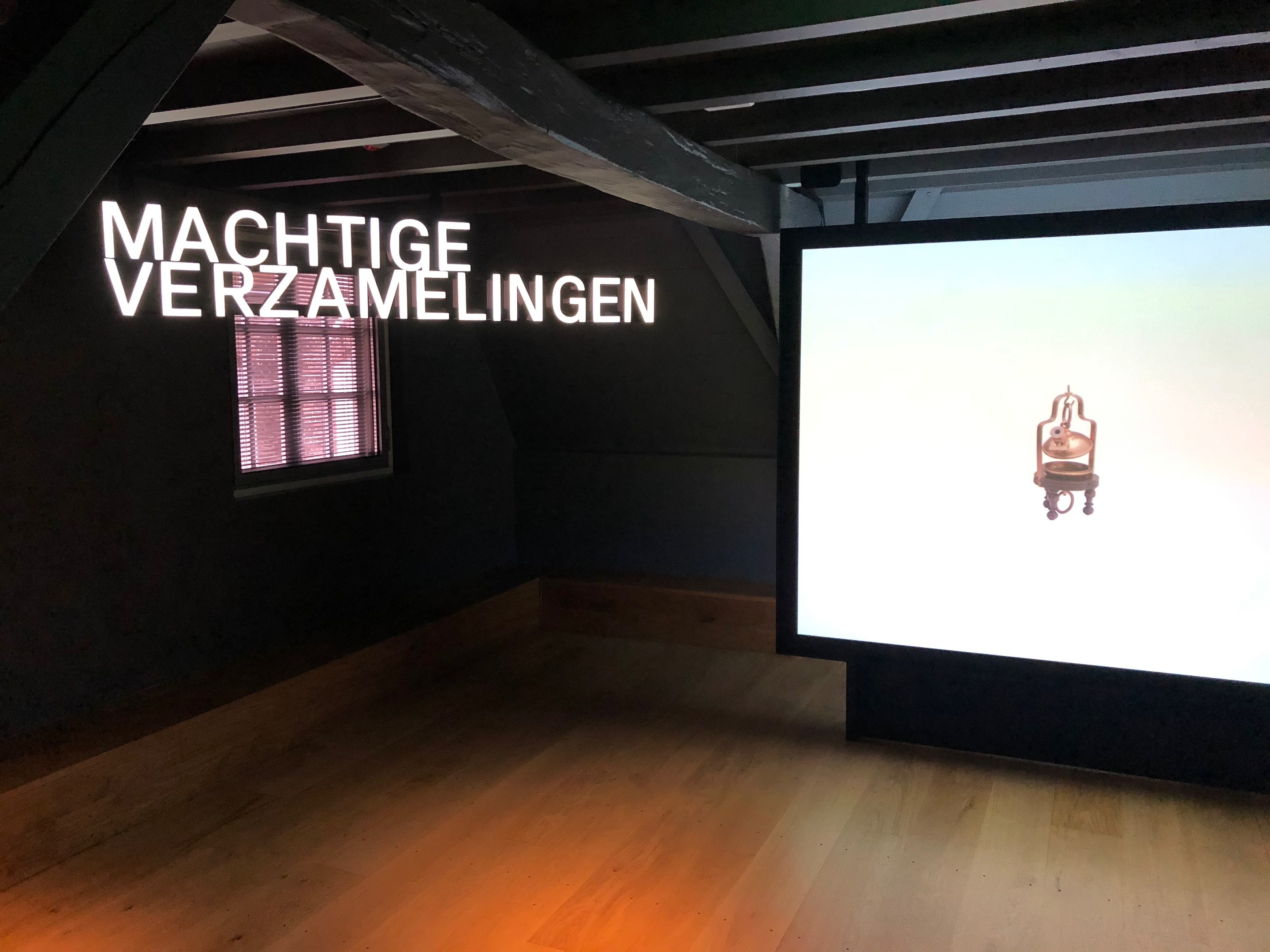 Installationsansicht der Ausstellung 'Machtige Verzamelingen' im Museum of Modern Art in Berlin, mit einem großen Bildschirm auf der rechten Seite und Text an der Wand dahinter sowie einem Fenster auf der linken Seite.