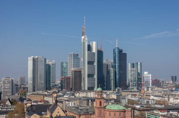 Der Frankfurter Skyline von einem Hügel aus betrachtet, mit einem prominenten Wolkenkratzer in der Mitte, umgeben von verschiedenen Gebäuden und Bäumen, unter einem klaren Himmel.