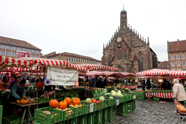 Ein belebter Markt in Nürnberg, Deutschland, mit verschiedenen Obst- und Gemüsesorten, Menschen mit Taschen, Zelten, Gebäuden mit Fenstern, einem Uhrenturm und einem sichtbaren Himmel.