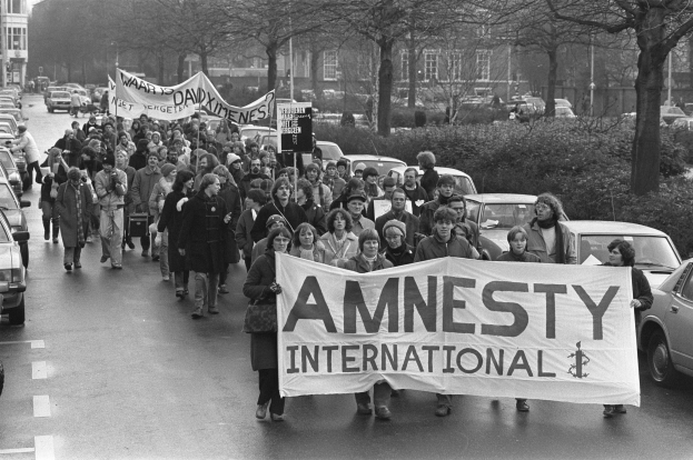 Eine Gruppe von Menschen marschiert mit einem Banner, auf dem 'Amnesty International' steht, die Straße entlang, mit parkenden Autos, Bäumen und Gebäuden im Hintergrund, dargestellt in Schwarz-Weiß.