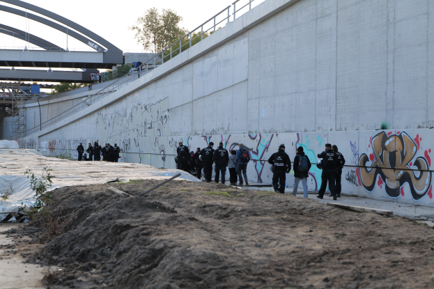 Gruppe von Polizisten in schwarzen Uniformen mit Taschen auf einem Schotterweg neben einer Wand mit Graffiti, mit einer Brücke, Bäumen und einem klaren blauen Himmel im Hintergrund.