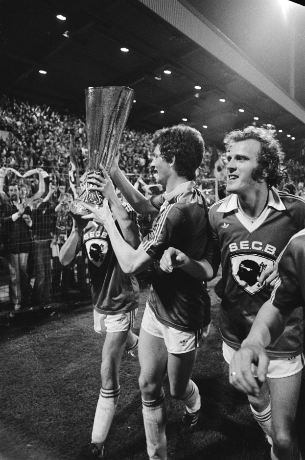 Schwarzes und weißes Foto von Fußballspielern, die mit einem Pokal auf einem Feld feiern, umgeben von einer Menge hinter einem Zaun unter Stadionlichtern.