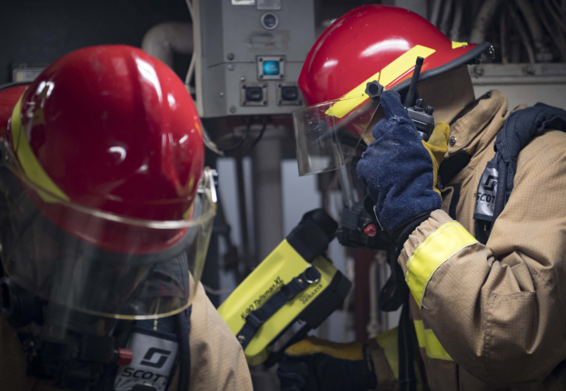 Zwei Feuerwehrleute in Schutzausrüstung bei der Arbeit an einem Feuerhydranten während einer Ausbildung mit Maschinen und Kabeln im Hintergrund.