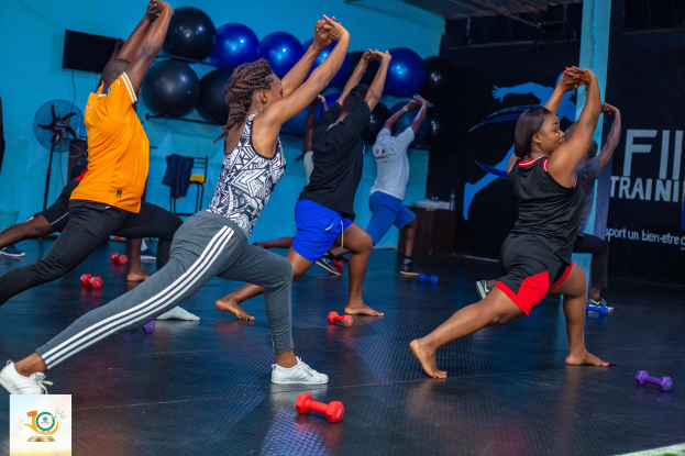 Gruppe von Menschen, die Yoga in einem Fitnessstudio mit Hanteln auf dem Boden machen, ein Banner mit der Aufschrift "Fitness-Training in Ghana" im Hintergrund.