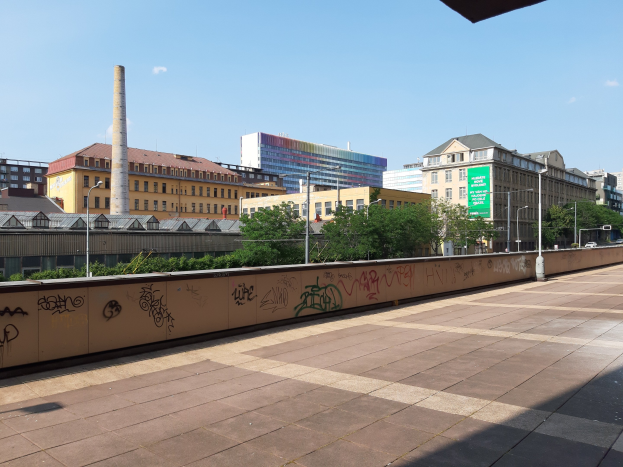 Stadtstraßenblick mit Graffiti-bedeckter Wand, Bäumen, Straßenlaternen, einem Schild, Fahrzeugen, Gebäuden mit Fenstern, einem Schornstein und einem bewölkten Himmel.