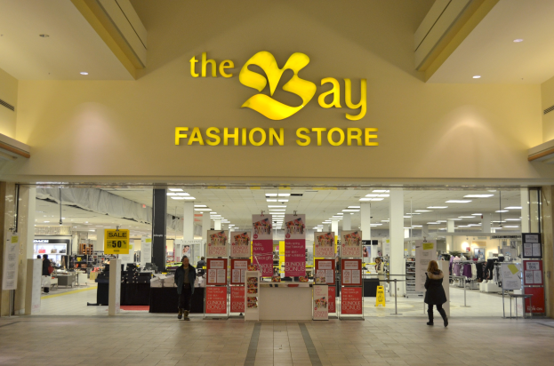 Außenansicht eines Bay Fashion Stores in einem Einkaufszentrum, das Menschen beim Gehen zeigt, Texttafeln an Wänden, Säulen, Deckenleuchten und Hintergrundbildschirme.