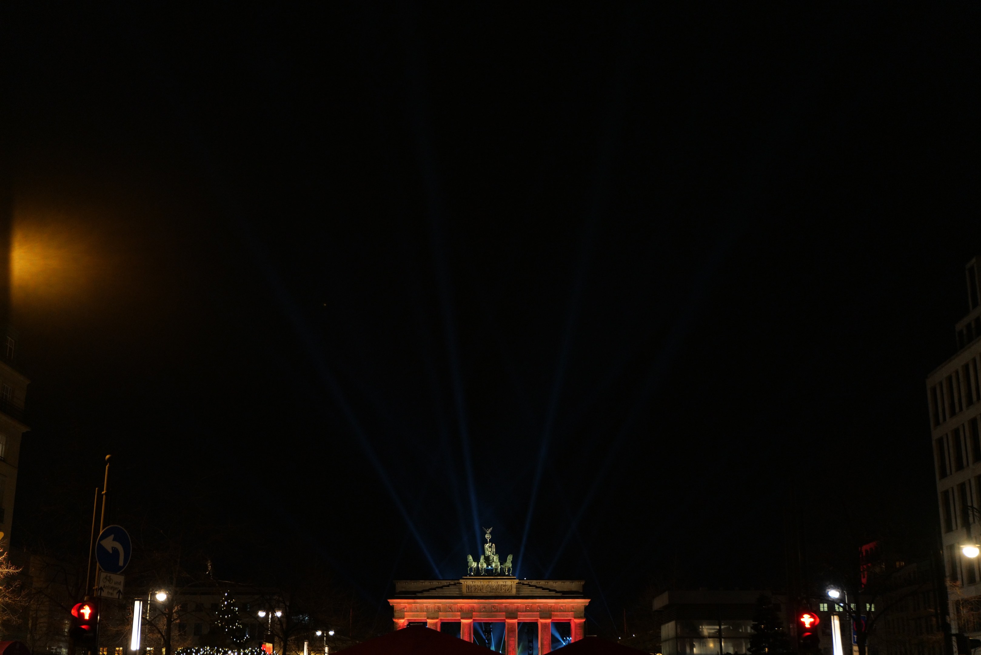 Brandenburger Tor in Berlin nachts, beleuchtet von Straßenlaternen, mit Gebäuden, Bäumen, Pfählen, Verkehrszeichen, Schildern und Zelten im Hintergrund, vor einem dunklen Himmel.