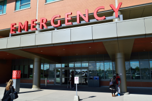 Eine Gruppe von Menschen geht vor einem Notfallgebäude mit Glaswänden, Säulen, einem Namensschild, Schildern auf Stangen und einem Mülleimer vorbei.