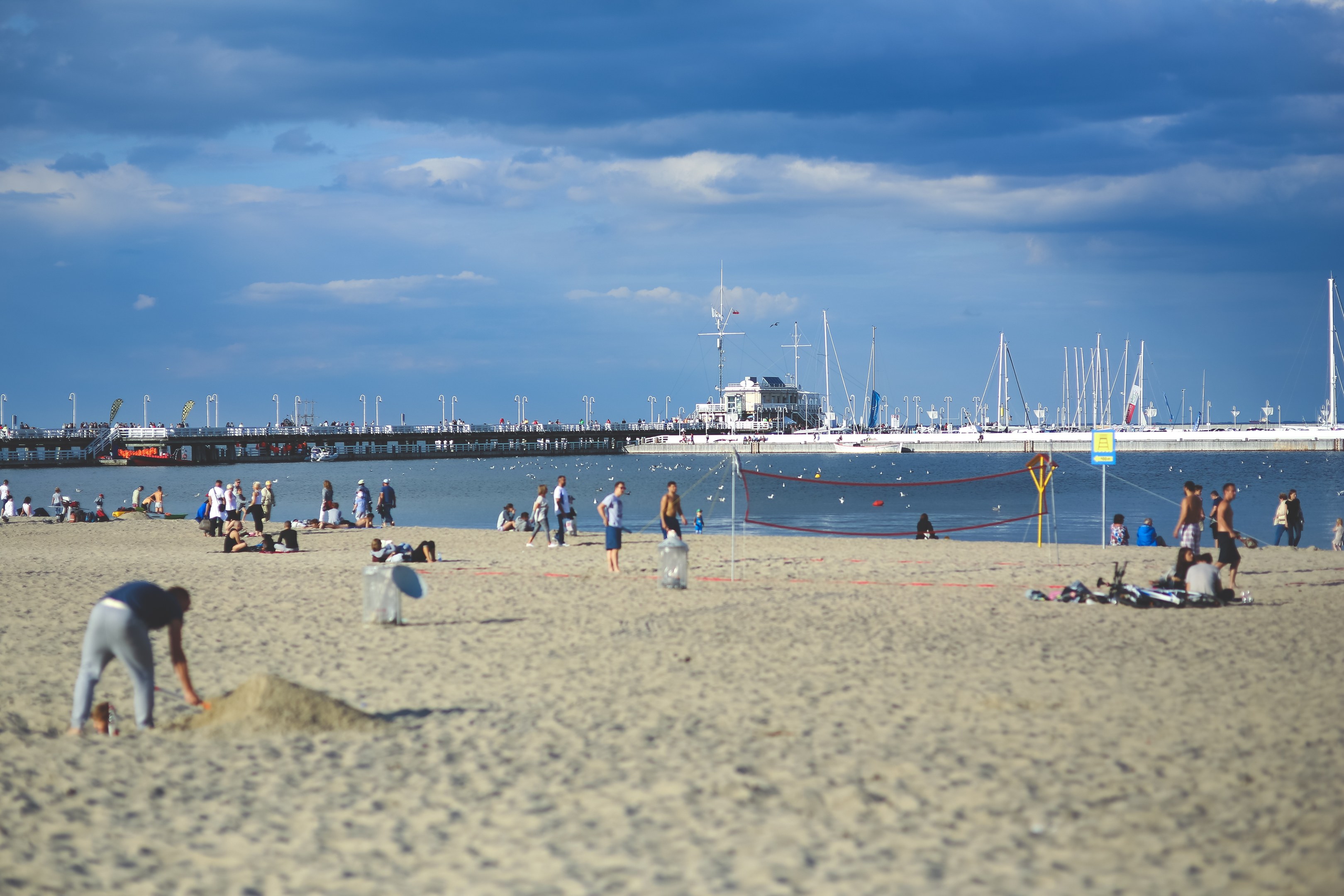 Menschen beim Volleyballspielen am Strand mit einem Netz, mit Booten und einer Brücke im Hintergrund bei einem bewölkten Himmel.