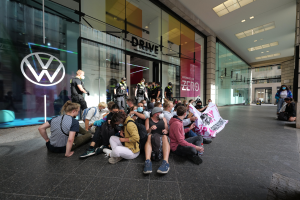 Gruppe von Menschen sitzt vor einem Volkswagen-Hndler auf dem Boden und halt eine Plakette waehrend einer Demonstration.