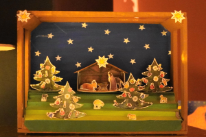 Eine Krippe in einer Holzkiste auf einem Tisch, mit einer Lampe links und einer Wand im Hintergrund, mit Dekorationen wie Weihnachtsbäumen, Tieren und einem Haus.