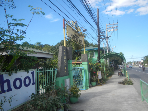 Der Eingang einer Schule mit einem Schild, auf dem "Schule" steht, auf der Seite einer Straße, mit einem Torbogen, einem Tor, einer Namensschild, Pflanzen, Bäumen, Strommasten mit Drähten und einem bewölkten Himmel im Hintergrund.