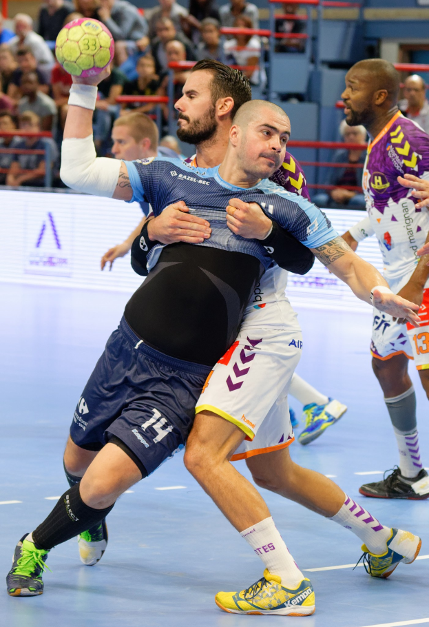 Eine Gruppe von Männern spielt Handball in einem Stadion, mit Zuschauern im Hintergrund.