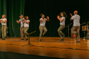 Eine Gruppe von Menschen auf einer Bühne mit Mikrofonen, Musikinstrumenten und einem Vorhang im Hintergrund, die Hände klappend, um ihren Erfolg zu feiern, scheinbar eine Tanzmannschaft.
