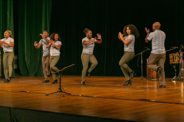Eine Gruppe von Menschen auf einer Bühne mit Mikrofonen, Musikinstrumenten und einem Vorhang im Hintergrund, die Hände klappend, um ihren Erfolg zu feiern, scheinbar eine Tanzmannschaft.