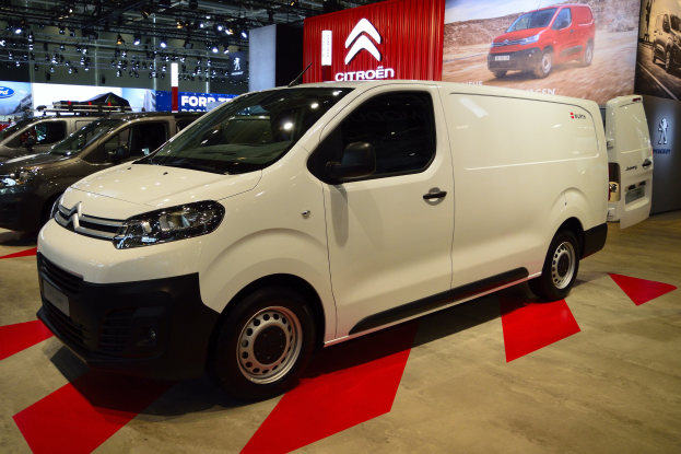 Ein weißer Citroen Berlingo Van wird auf einer Autauschau präsentiert, umgeben von schwarzer Schlafzimmer-Möblierung, mit Wänden, die mit Brettern und Lichtern geschmückt sind.