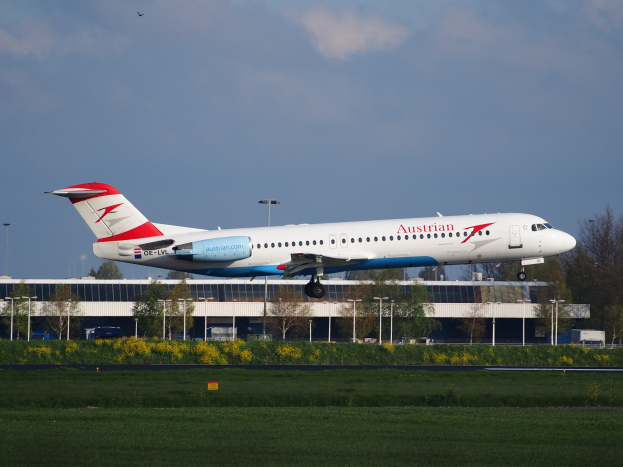 Ein Austrian-Airlines-Airbus-A320-200 beim Starten am Frankfurter Flughafen mit Grünflächen und einem Gebäude im Hintergrund bei einem teilweise bewölkten Himmel.