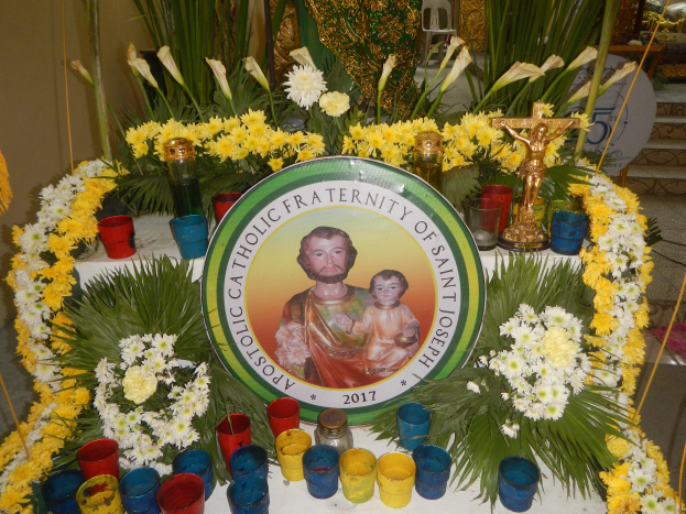 Ein mit bunten Blumen, Kerzen und einer Tafel mit einem Porträt und Text geschmückter Tisch vor einer Kulisse aus Pflanzen, Stühlen und anderen Gegenständen schafft eine festliche Atmosphäre für die Bruderschaft des Heiligen Josef.