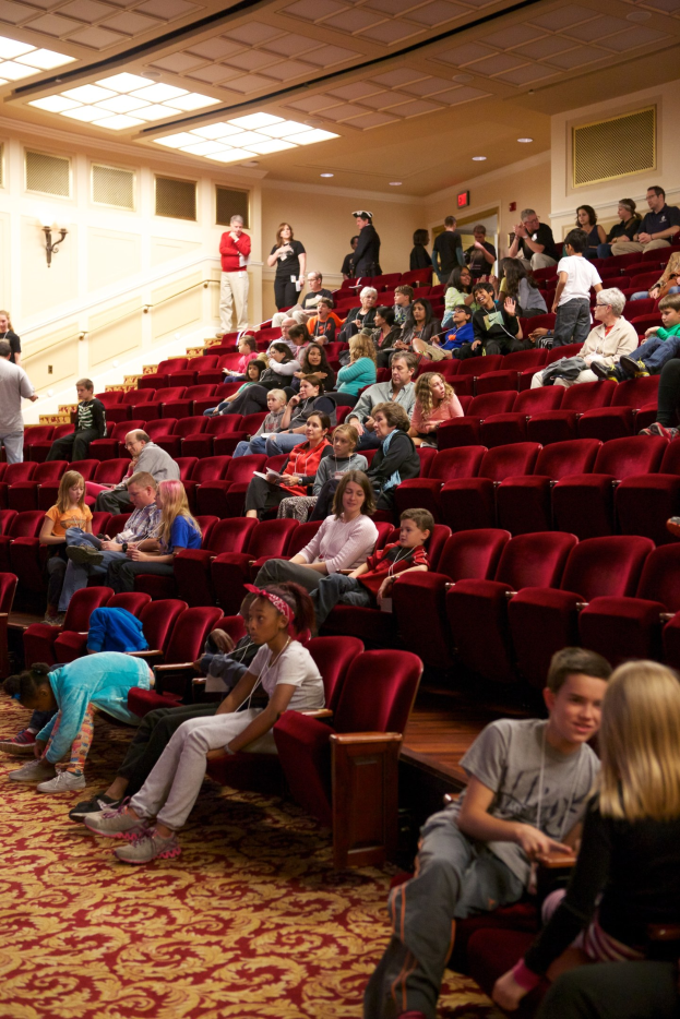 Eine große Gruppe von Menschen, die auf Stühlen sitzen oder stehen, in einem Theater mit verstreuten Stühlen, einer beleuchteten Wand im Hintergrund und Kindern in der Mitte.
