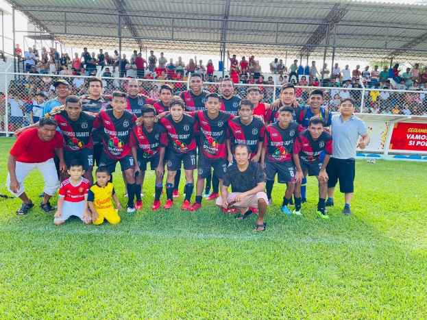 Gruppe von Männern, die auf einem Fußballfeld mit einem Banner feiern, das "Fotos Ugarte U-17 vs U-18" trägt, und einem Metallzaun.