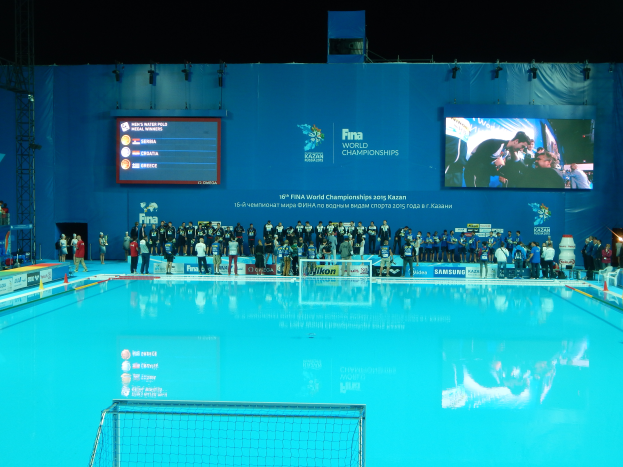 Eine Gruppe von Menschen um einen Swimmingpool mit Netz am Boden herumstehend, mit einem Bildschirm im Hintergrund, der "FINA World Championships 2015 Kazan" sowie Logos, Träger und Lichter anzeigt.