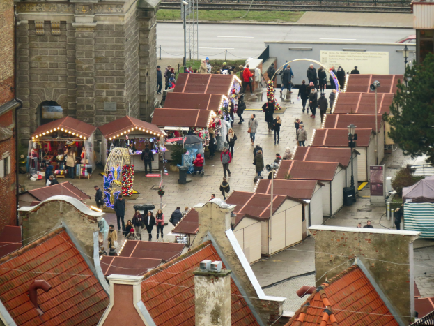 Eine belebte Stadtstraße mit zahlreichen Fußgängern, Gebäuden, Ständen, einem Baum, einem Schild, einer Bahnschiene und einem bewölkten Himmel.