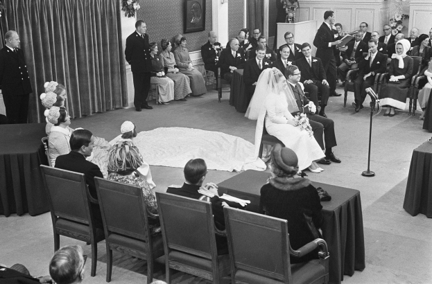 Schwarze und weiße Fotografie einer Hochzeitszeremonie mit sitzenden Gästen, einem Mann mit Mikrofon, einer Frau mit Blumenstrauß, einem Podium mit Mikrofon, Blumenschmuck, einem Vorhang, einem gerahmten Foto an der Wand und einer Tür im Hintergrund.