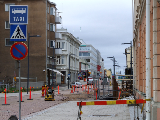 Stadtstraße mit Gebäuden, Straßeninfrastruktur, Fahrzeugen, einer Baustelle und Bäumen unter einem bewölkten Himmel.