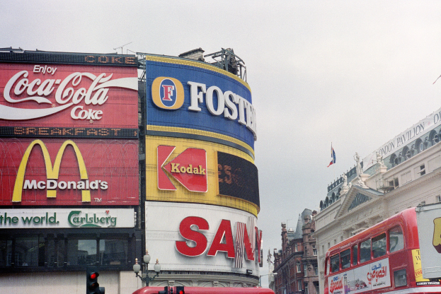 Ein roter Doppeldeckerbus fährt auf einer belebten Straße mit hohen Gebäuden, Ampeln, Laternenmasten und Texttafeln, vor einem Himmel mit einer Flagge auf einem Gebäude im Hintergrund.