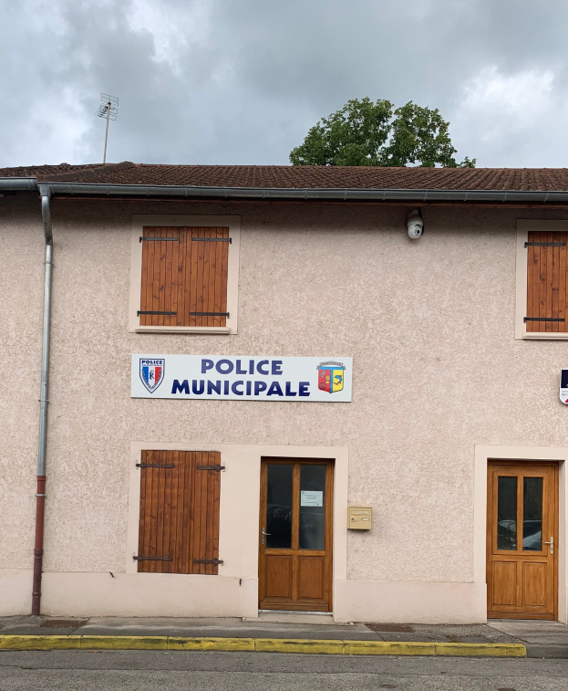 Ein Gebäude der police municipale mit einem Schild, Fenstern, Türen, einer Namensplatte, einem Rohr, einem Baum und einem bewölkten Himmel im Hintergrund.
