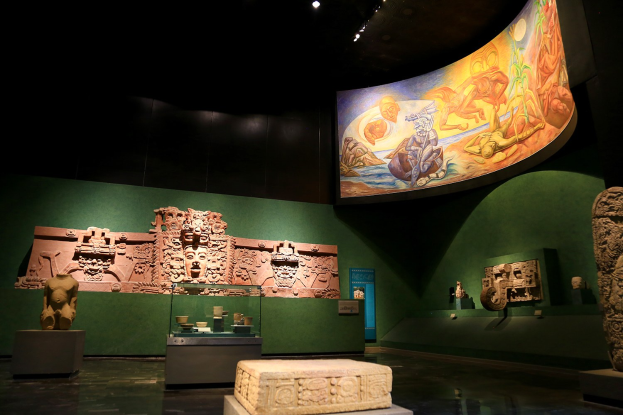 Ein Museumsraum mit verschiedenen Kunstwerken, darunter Skulpturen, Objekte auf Tischen, eine Texttafel, eine Wandmalerei und Deckenlampen im Mexiko-Stadt-Museum der Anthropologie.