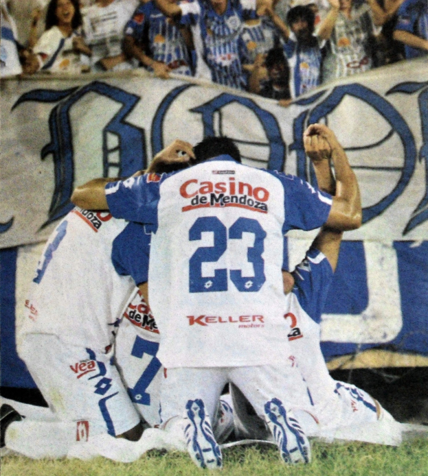 Gruppe von Fussballspielern feiert auf dem Boden vor einer jubelnden Menge, mit einem Banner mit Text im Hintergrund.