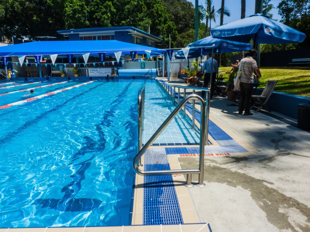 Großer Freibadpool mit schwimmenden Menschen, umgeben von Geländern, Sonnenschirmen, Stühlen, Tischen, Pflanzen, Gras, Bäumen und einem Gebäude, unter einem klaren blauen Himmel.