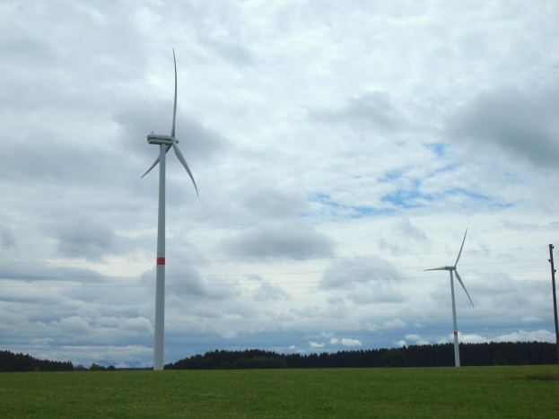 Drei hohe weiße Windräder auf einem grünen Feld mit Bäumen und Wolken im Hintergrund.