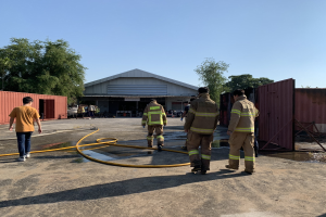Eine Gruppe von Feuerwehrleuten in Uniformen steht vor einem Gebäude mit einem Schlauch, umgeben von Containern, Bäumen und anderen Gegenständen, mit Wasser auf der rechten Seite und dem Himmel im Hintergrund.