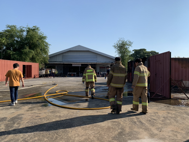 Eine Gruppe von Feuerwehrleuten in Uniformen steht vor einem Gebäude mit einem Schlauch, umgeben von Containern, Bäumen und anderen Gegenständen, mit Wasser auf der rechten Seite und dem Himmel im Hintergrund.