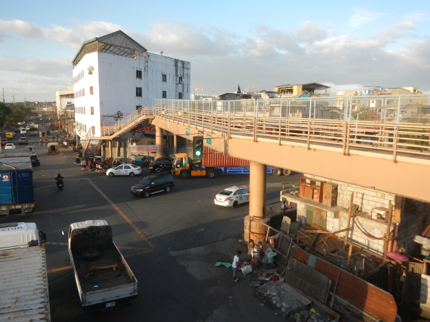 Eine belebte Stadtstraße mit Autos, Lastwagen und Fußgängern, eine Brücke mit Geländern und Pfeilern, Ampeln, Gebäude mit Fenstern, Bäume und ein bewölkter Himmel.