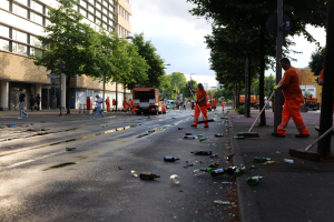 Eine Gruppe von Menschen in orangefarbenen Uniformen, die Müll von einer Straße mit Flaschen und Schutt säubern, mit Bäumen, Pfählen und Fahrzeugen im Hintergrund unter einem bewölkten Himmel.
