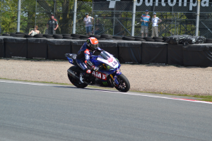 Ein Motorradfahrer in Helm und Handschuhen fährt auf einer von Gras, Reifen, Maschendrahtzaun mit Pfosten und einer Fahne umgebenen Rennstrecke, mit Bäumen und einem klaren blauen Himmel im Hintergrund.