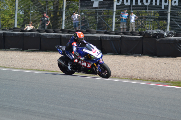 Ein Motorradfahrer in Helm und Handschuhen fährt auf einer von Gras, Reifen, Maschendrahtzaun mit Pfosten und einer Fahne umgebenen Rennstrecke, mit Bäumen und einem klaren blauen Himmel im Hintergrund.