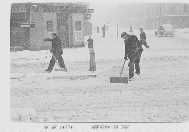 Ein Schwarz-Weiß-Foto von Menschen, die Schnee schippen, auf einer Stadtstraße mit Gebäuden, Pfählen, Brettern und Fahrzeugen im Hintergrund und Text am unteren Rand des Bildes.
