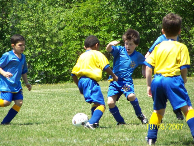 Kinder beim Fußballspielen auf einer Wiese mit Bäumen im Hintergrund und einem Datumsstempel in der rechten unteren Ecke.
