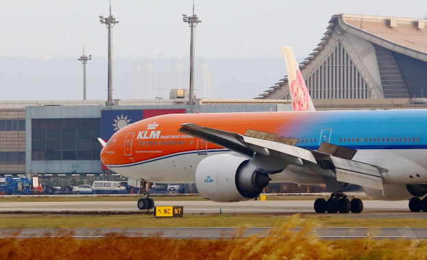 KLM Boeing 777-300ER Flugzeug auf der Rollbahn am Frankfurter Flughafen mit Gras und Pflanzen im Vordergrund, Gebäuden und Fahrzeugen im Hintergrund und einem klaren blauen Himmel.