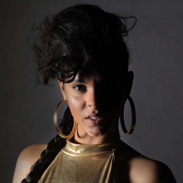 Eine Frau in einem goldenen Kleid und großen Ohrringen steht selbstbewusst vor einer Wand, ihre glatten Bob-Haare ordentlich frisiert, dezent natürliches Make-up mit pinkem Rouge und glänzenden Lippen, blickt direkt in die Kamera mit einem entschlossenen Gesichtsausdruck.